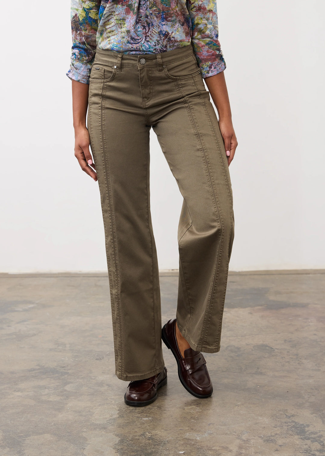 A model wearing a pair of olive green wide-leg pants, styled with a colorful floral long-sleeve shirt and dark brown leather loafers, standing against a plain white wall and concrete floor.
