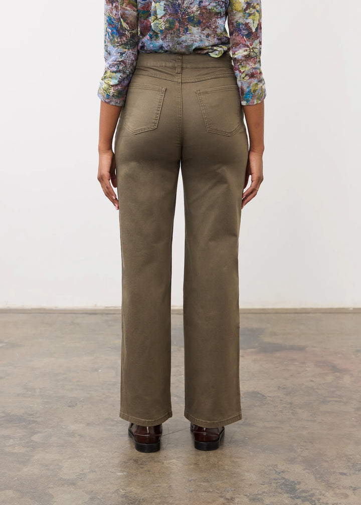A person shown from the back wearing olive brown wide-leg trousers, a colorful floral long-sleeve top, and dark brown shoes, standing against a plain white wall and concrete floor.
