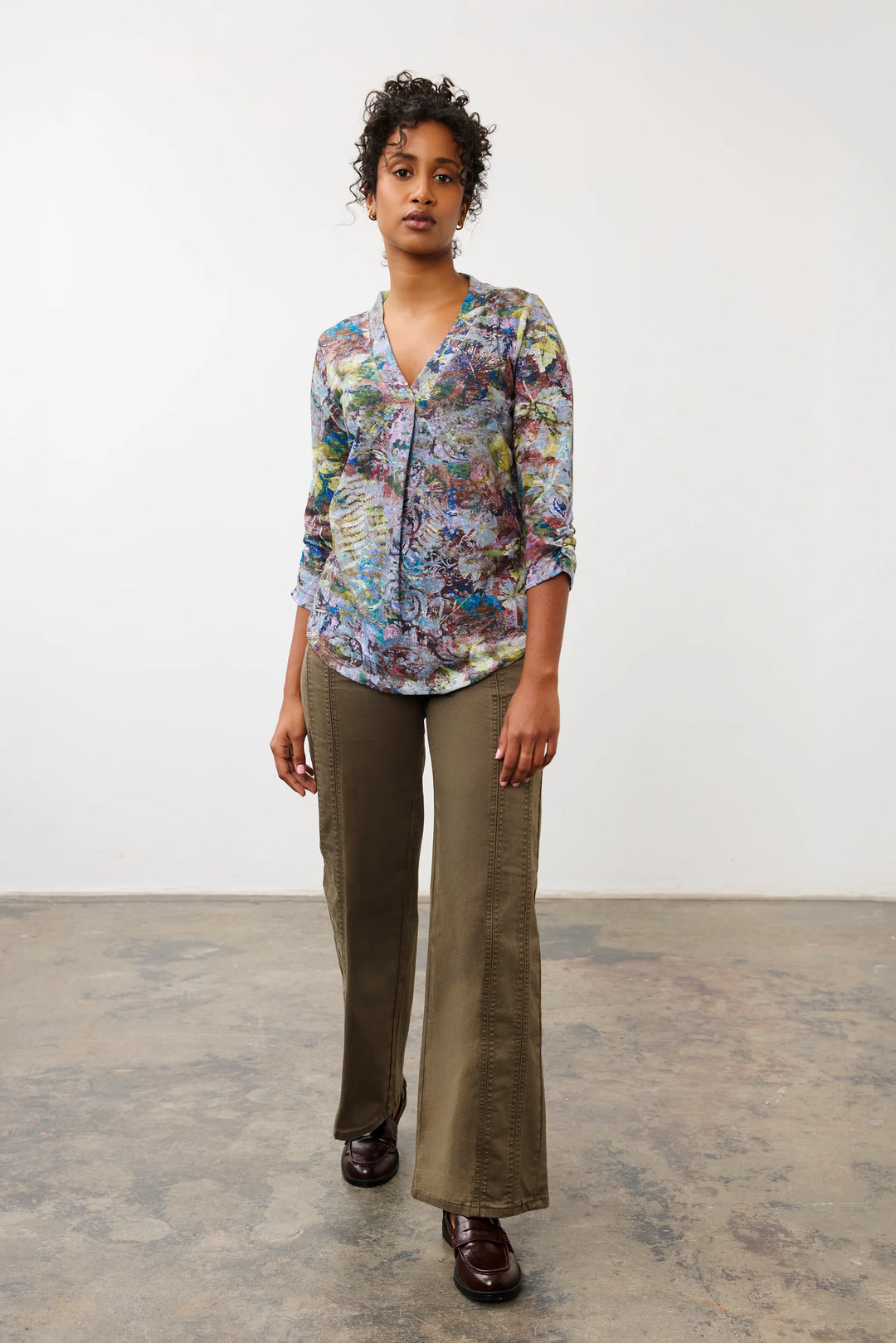 A model stands in a studio wearing a colorful abstract print button-up shirt with olive green wide-leg pants and dark brown leather shoes.