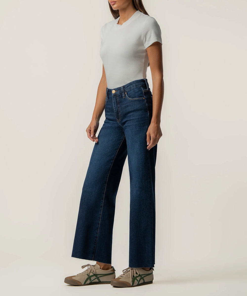 A model wearing dark blue wide-leg jeans, a light gray short-sleeve top, and brown sneakers with green stripes, standing against a plain off-white studio background.