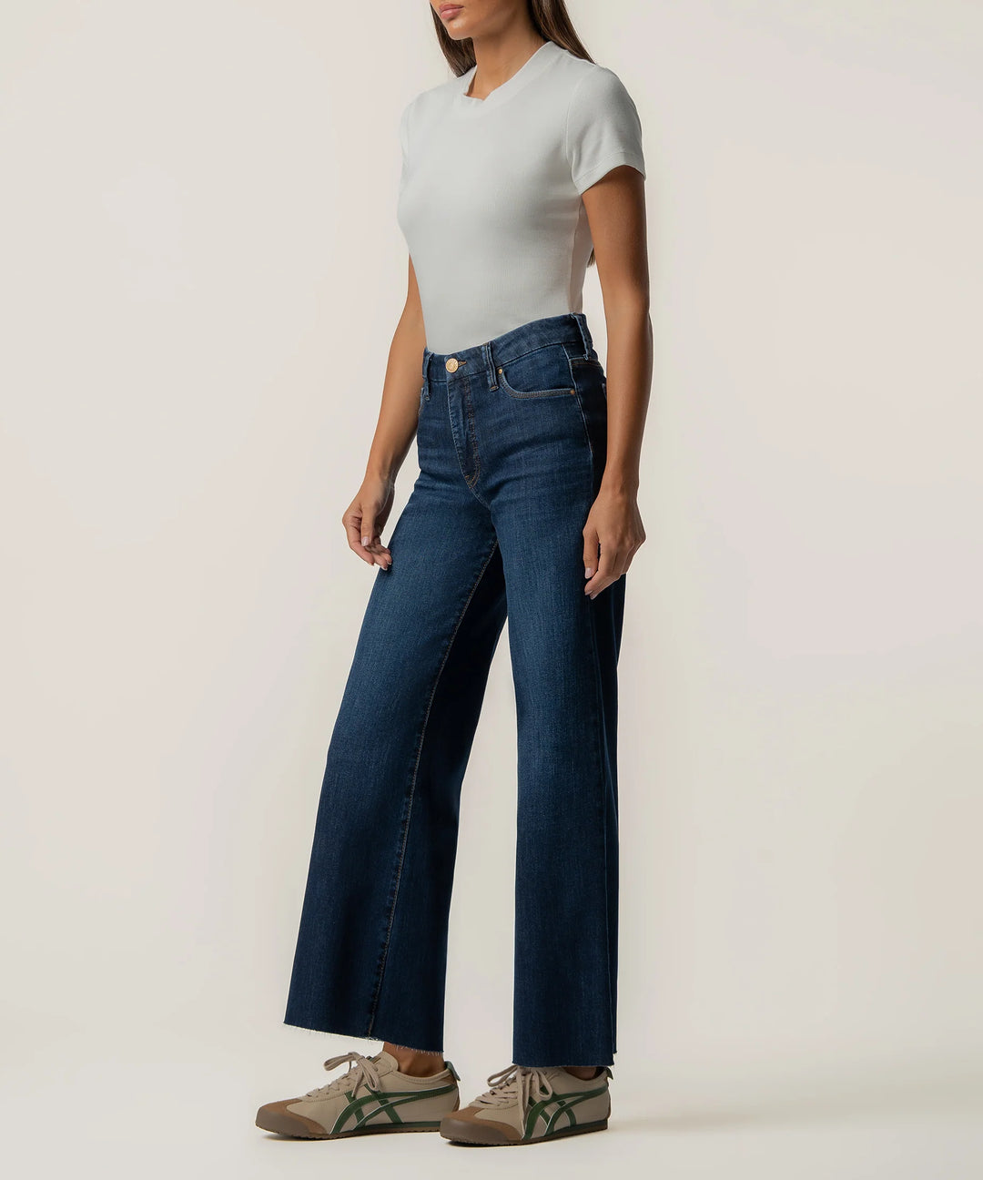 A model wearing dark blue wide-leg jeans, a light gray short-sleeve top, and brown sneakers with green stripes, standing against a plain off-white studio background.