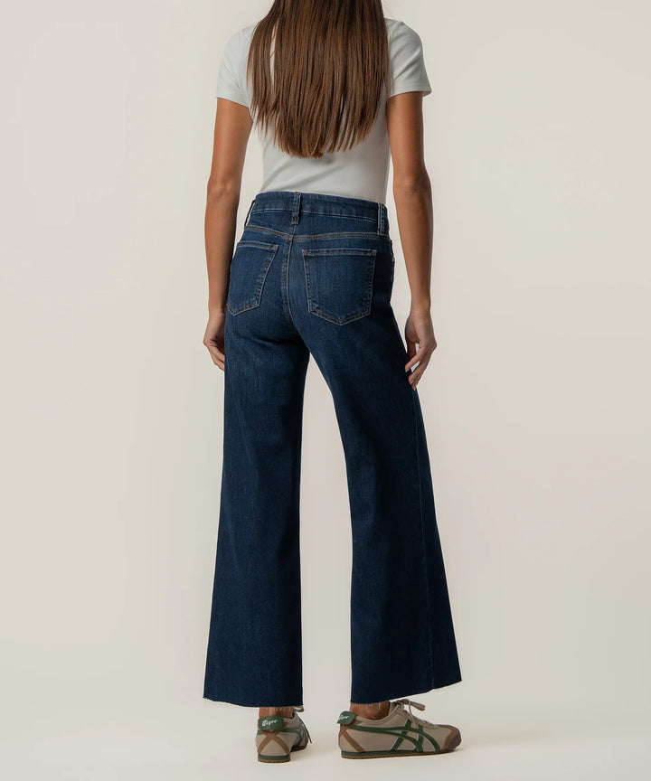A woman shown from the back wearing dark blue wide-leg flare jeans, a light gray t-shirt, and green-and-brown slide sandals against a plain off-white studio background.