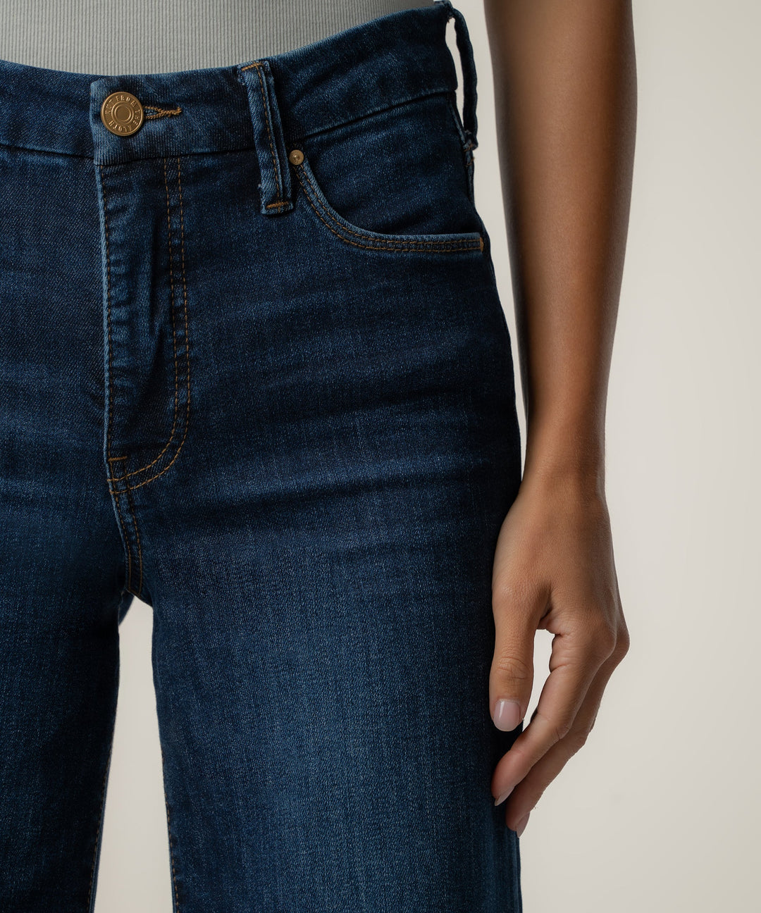 Close-up of a person wearing dark blue denim jeans, showing the front waistband, belt loops, pocket, and hand against a plain light background.