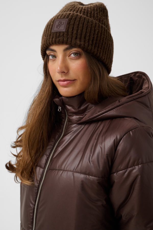 A woman wearing a brown ribbed knit beanie with a small square logo patch and a shiny dark brown hooded puffer jacket, photographed against a light gray studio background.