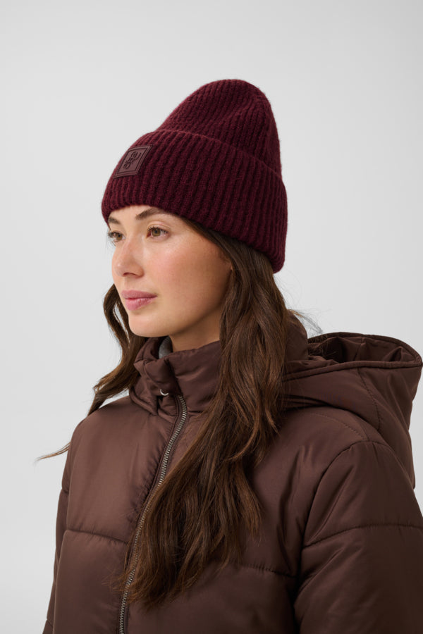 A woman wearing a dark burgundy ribbed knit beanie and a brown puffer jacket, shown in profile against a light gray studio background.