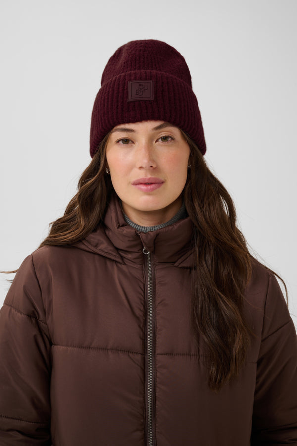 A woman wearing a dark burgundy ribbed knit beanie with a small square logo patch and a brown puffer jacket, photographed against a light gray studio background.