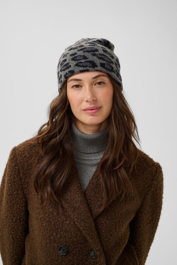 A woman wearing a gray and black leopard-print knit beanie, a gray turtleneck sweater, and a brown textured fleece coat, photographed against a light gray studio background.