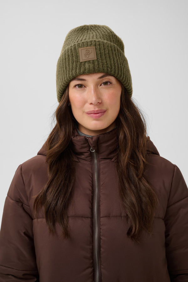 A woman wearing an olive green ribbed knit beanie with a small brown brand patch and a brown puffer jacket, photographed against a light gray studio background.