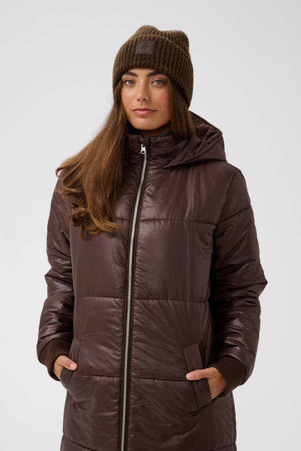 A woman wearing a dark brown hooded puffer jacket with a front zipper, matching brown knit beanie, and brown knit gloves, photographed against a light gray studio background.