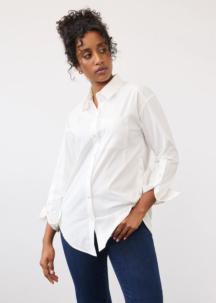 A woman wearing a white long-sleeve button-up shirt with rolled cuffs and dark blue jeans, photographed against a plain light gray studio background.