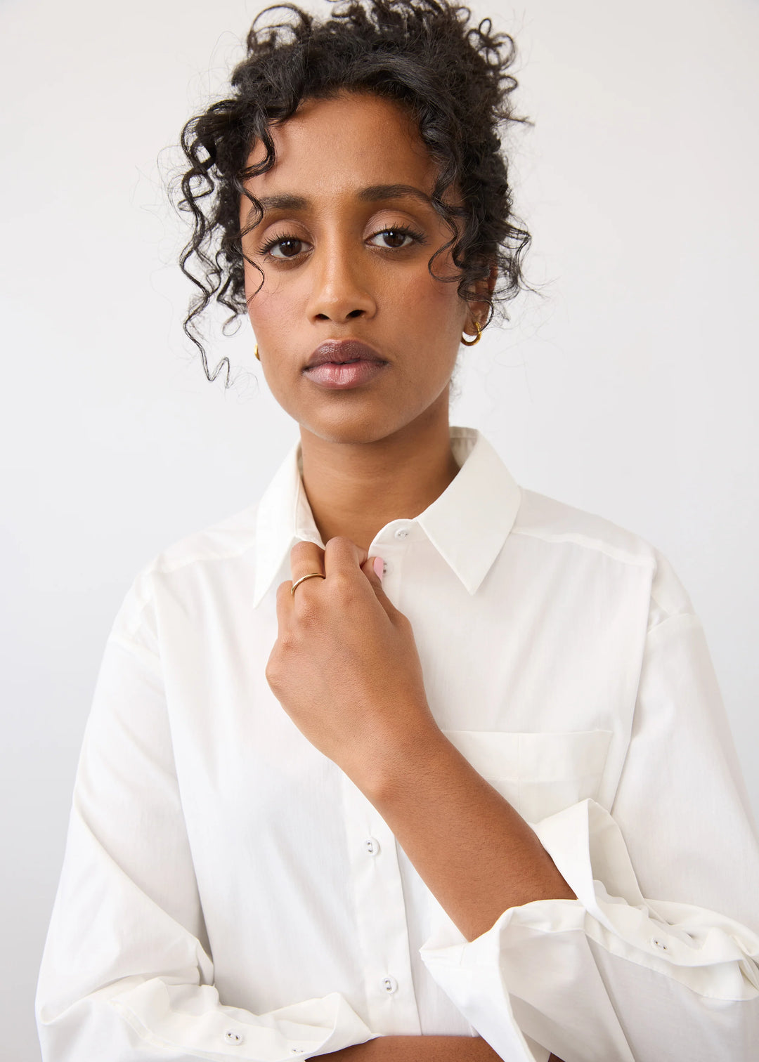 A woman with curly dark hair wearing a white long-sleeve button-up shirt, posing against a plain light background.