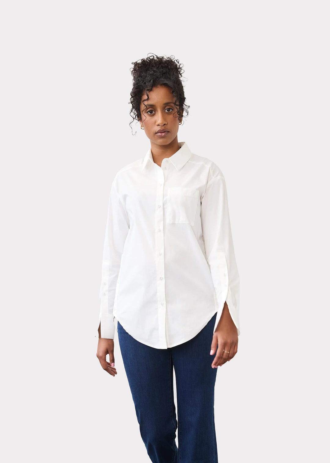 A model stands in a studio wearing a white long-sleeve button-up shirt, dark blue cropped pants, and black pointed flats against a light gray wall and concrete floor.
