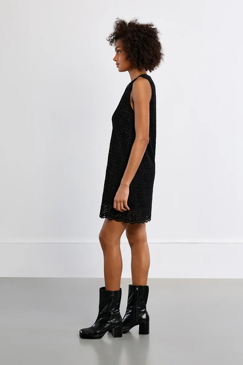 A woman stands in profile wearing a sleeveless black textured mini dress and black ankle boots against a clean white studio wall and gray floor.