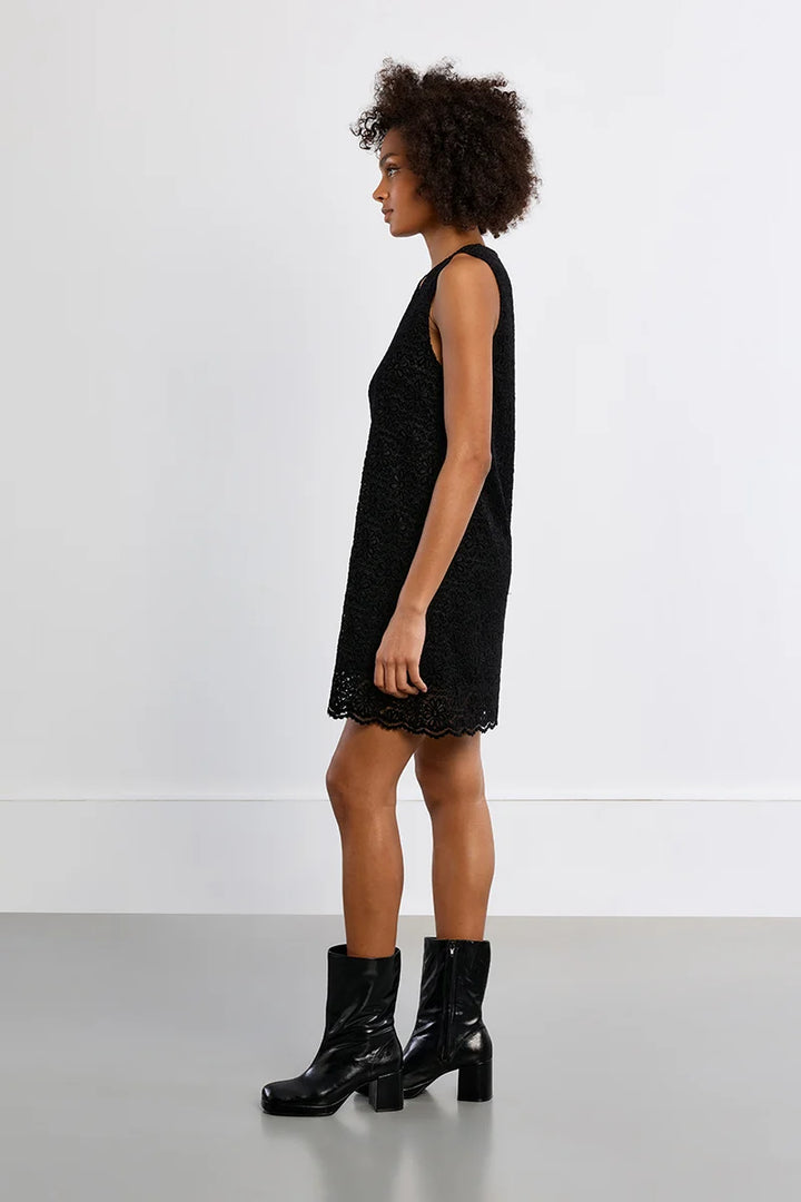 A woman stands in profile wearing a sleeveless black textured mini dress and black ankle boots against a clean white studio wall and gray floor.