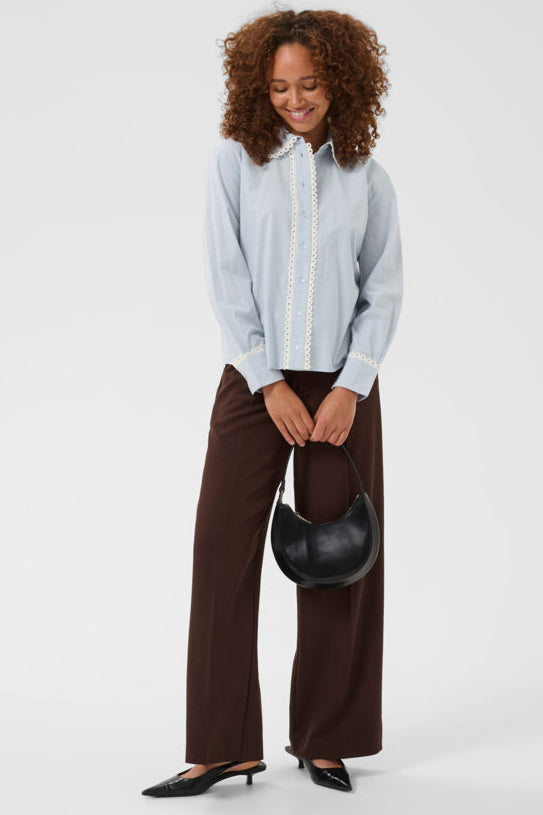 Woman wearing a light blue shirt and brown pants holding a black handbag on a white background