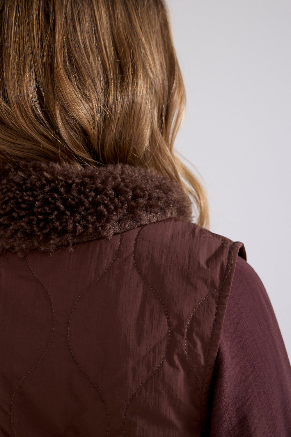 Close-up back view of a person wearing a dark brown quilted vest with a thick dark brown faux-fur collar, paired with a mauve-purple long-sleeve shirt, against a light gray background.