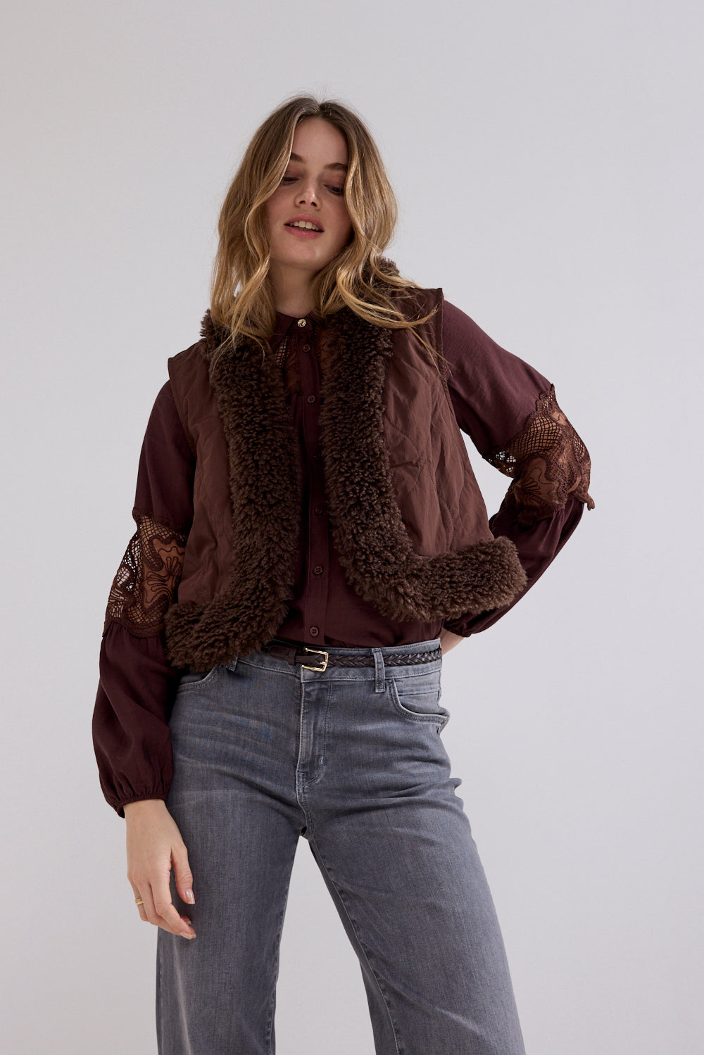 A woman modeling a dark brown cropped jacket with a brown faux-fur collar and lace-up sleeves, paired with a matching brown blouse and gray jeans, against a light gray studio background.