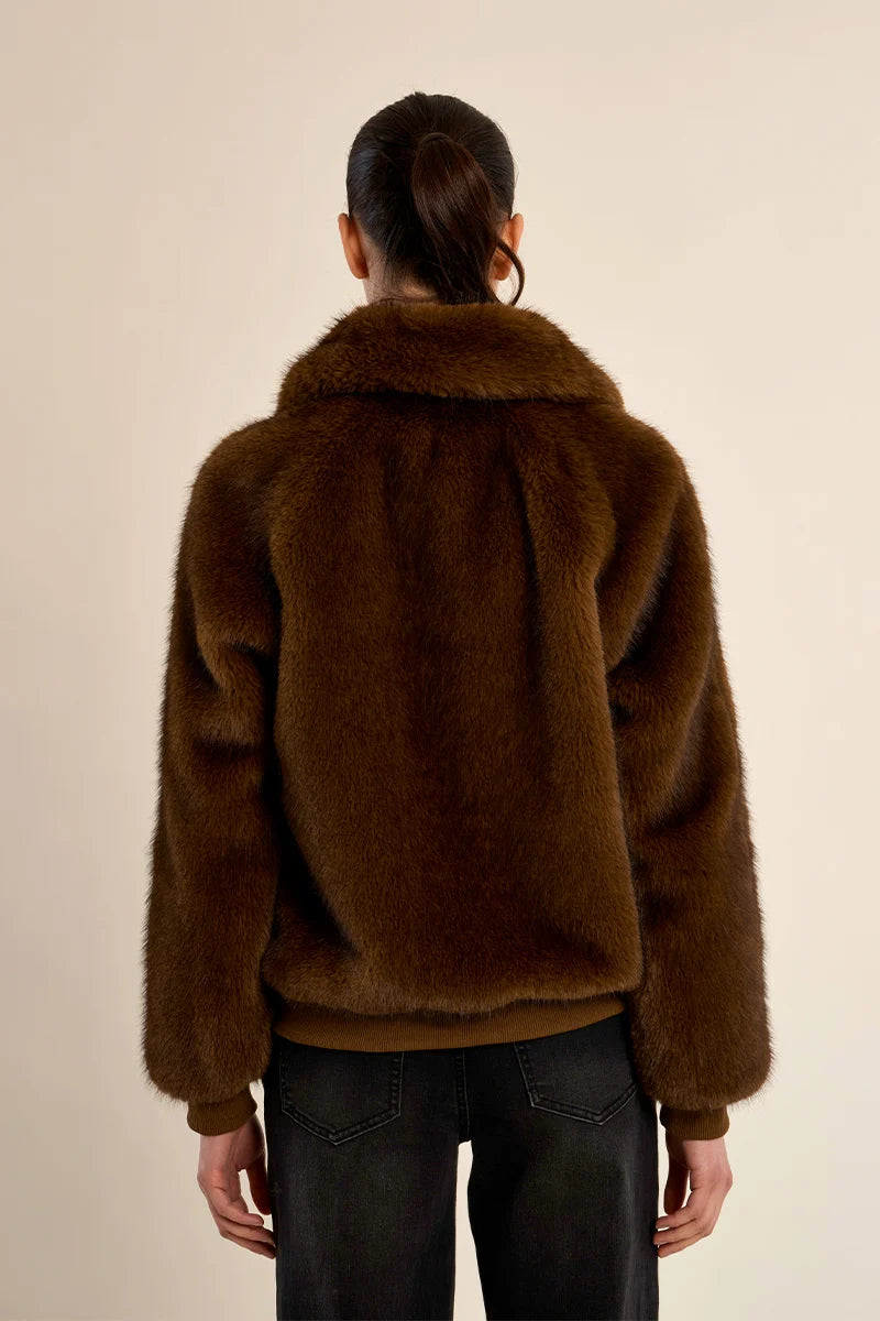 A person viewed from the back wearing a dark brown plush faux fur jacket with a wide collar, paired with black jeans, against a plain beige background.