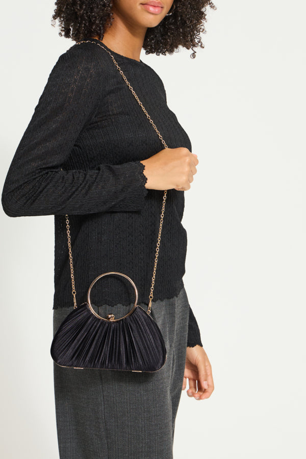 A model wearing a black ribbed knit sweater and gray trousers carries a black pleated clutch bag with a gold-tone circular ring handle and chain strap.