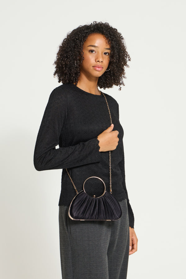A woman with curly dark hair models a black ribbed long-sleeve top and dark gray trousers, carrying a small black pleated shoulder bag with a gold-tone chain strap and circular ring detail against a light gray studio background.