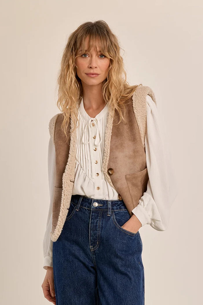 A woman modeling a sleeveless taupe faux-suede vest with cream sherpa lining over a cream button-front blouse and blue jeans, photographed against a plain warm beige studio background.