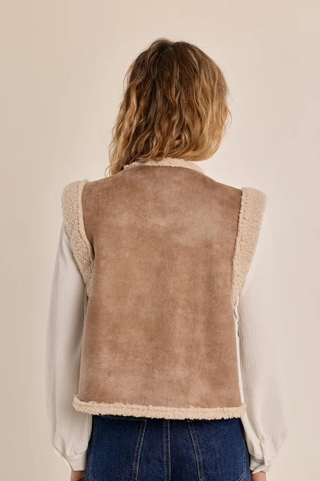 A woman viewed from behind wearing a sleeveless taupe-brown suede vest with cream sherpa lining and trim, layered over a white long-sleeve blouse and blue jeans, against a plain beige studio background.