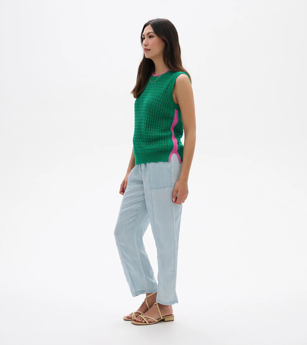 Woman wearing a green sleeveless top with pink accents and light blue pants on a white background
