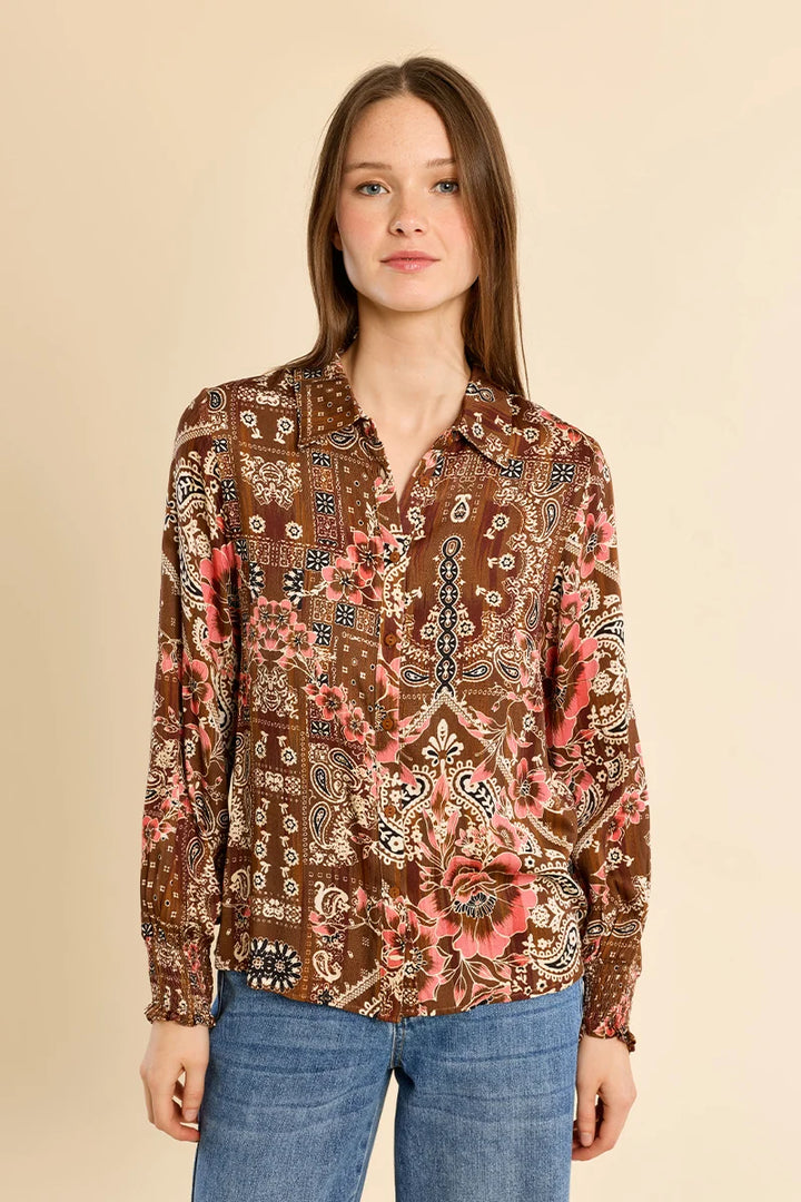 A woman modeling a long-sleeve button-up blouse with an intricate brown, pink, and beige paisley and floral pattern, paired with blue jeans against a solid beige background.