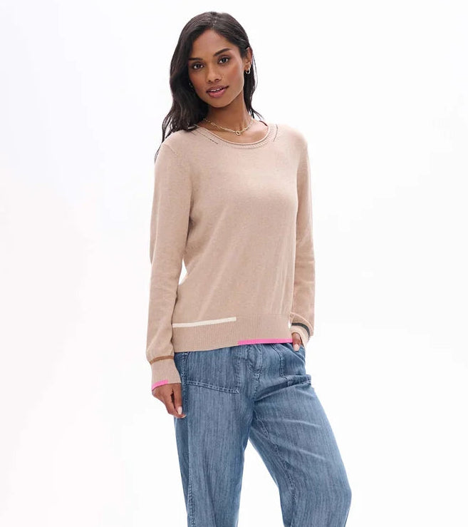 A woman modeling a light beige crewneck sweater with contrasting pink and white ribbed trim at the hem and cuffs, paired with blue jeans against a plain white background.