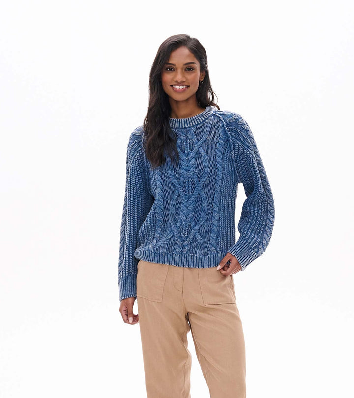 Woman wearing a blue cable knit sweater, beige pants, and brown boots on a white background