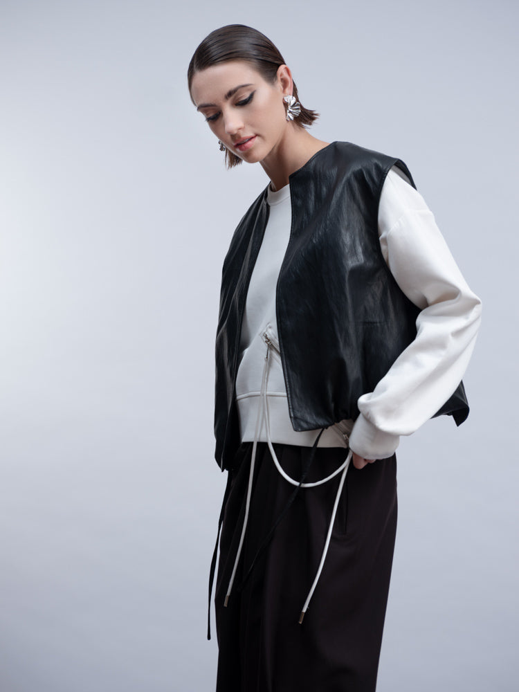A model wearing a black leather-look sleeveless vest over a white long-sleeve top and black drawstring pants, photographed against a light gray studio background.