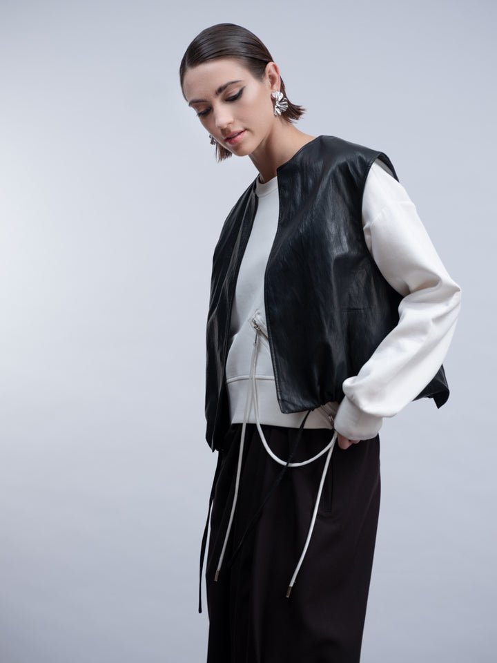 A model wearing a black leather-look sleeveless vest over a white long-sleeve top and black drawstring pants, photographed against a light gray studio background.
