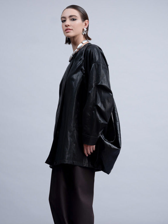 A model is shown in profile wearing a black oversized shiny jacket and dark wide-leg trousers against a light gray studio background.