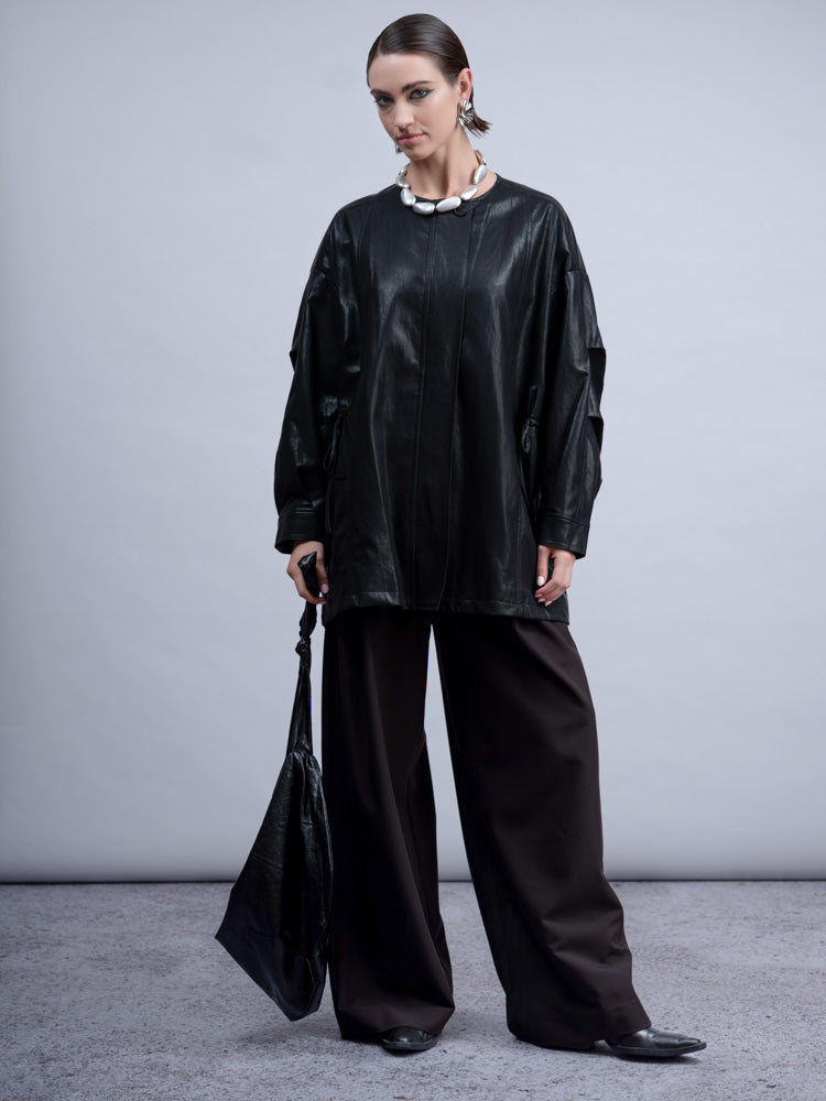 A model stands in a studio wearing an oversized black faux-leather top with draped sleeves, wide-leg dark trousers, a chunky white collar necklace, and black pointed shoes, while holding a black tote bag.