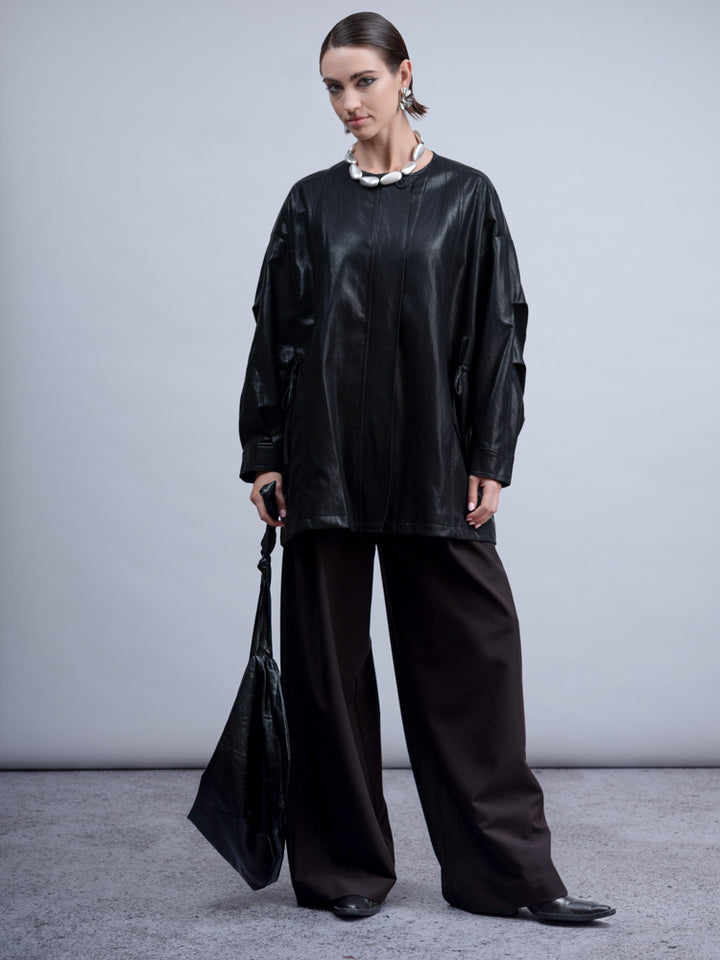 A model stands in a studio wearing an oversized black faux-leather top with draped sleeves, wide-leg dark trousers, a chunky white collar necklace, and black pointed shoes, while holding a black tote bag.