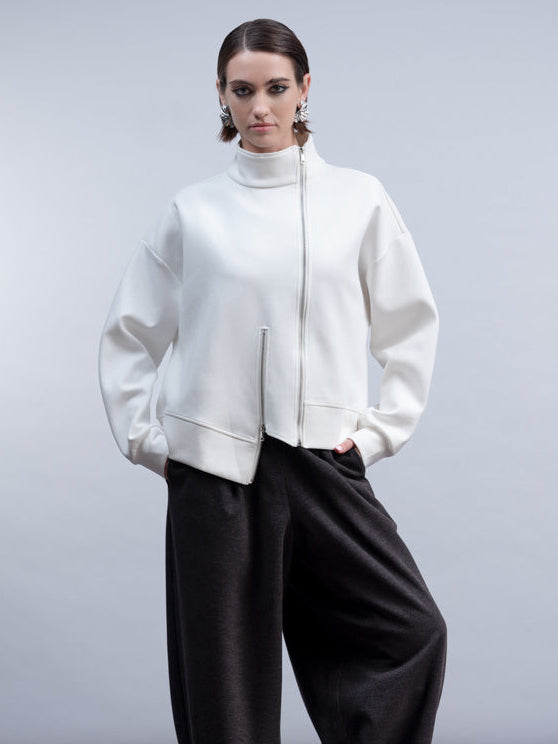 A model stands against a light gray studio background wearing a white cropped zip-front jacket with a high mock neck, black wide-leg pants, and black ankle boots.