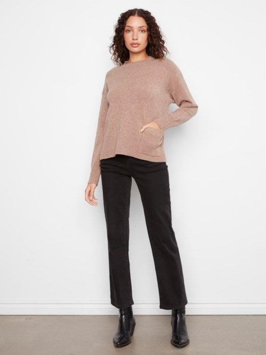 A woman modeling a light brown knit sweater with black pants and black ankle boots, standing against a white wall.