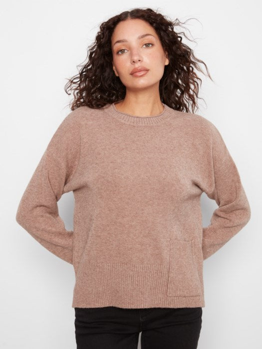 A woman with curly brown hair wearing a dusty rose knit crewneck sweater and black pants, standing against a light gray studio background.