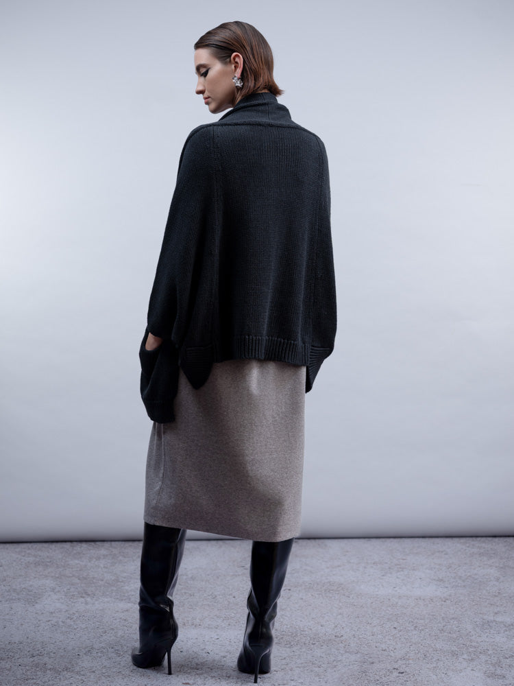 A model shown from the back wearing a black knit cape cardigan over a taupe knit midi skirt, paired with black knee-high boots, against a neutral gray studio background.