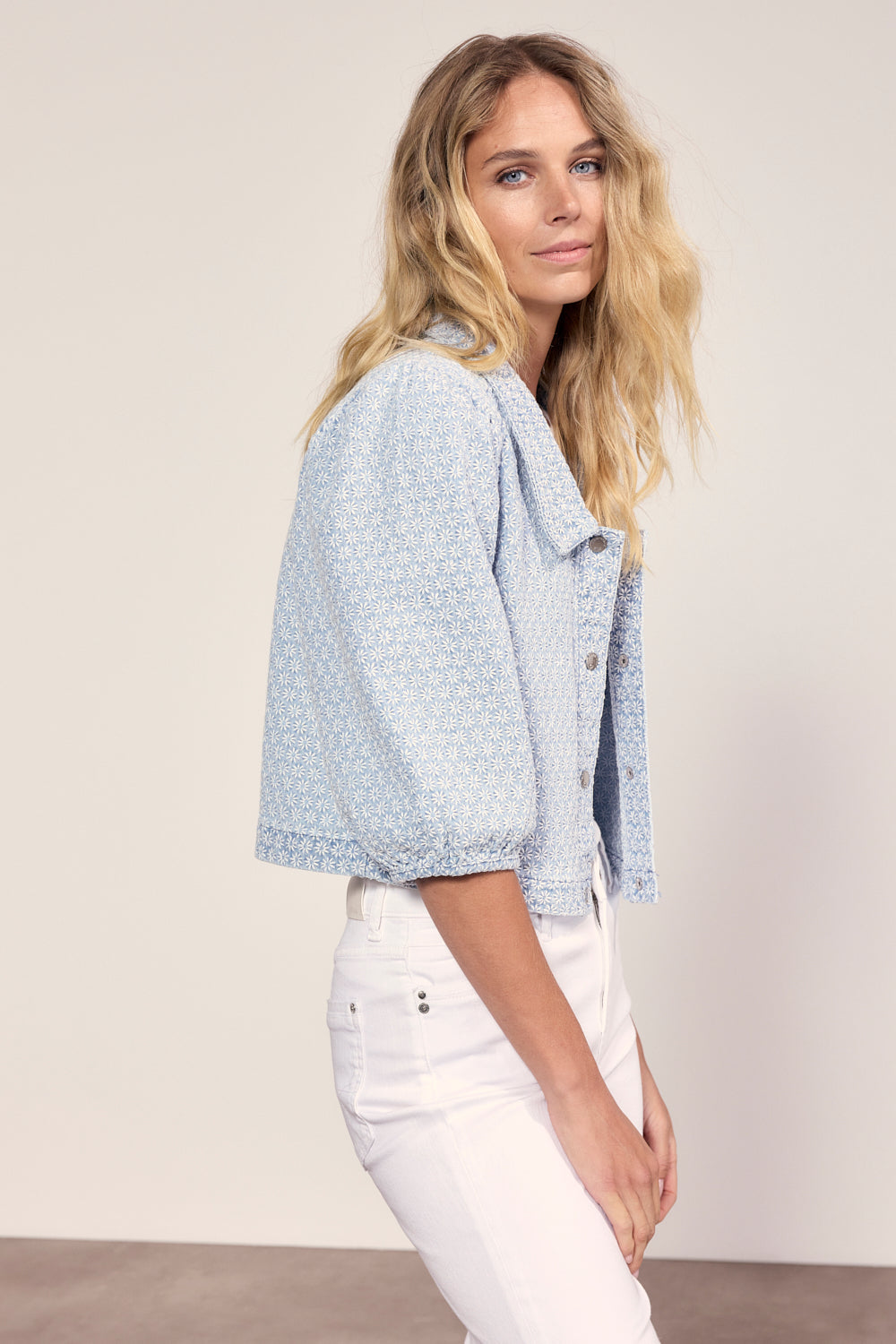 Woman wearing a light blue patterned jacket and white pants against a plain background