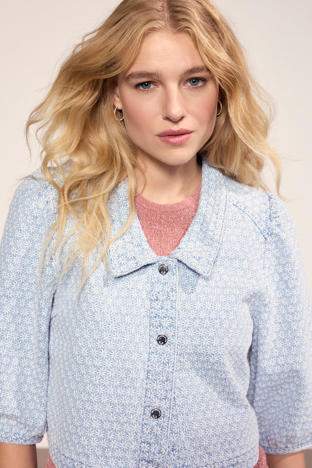 Woman wearing a light blue patterned shirt with a pink top underneath against a plain background