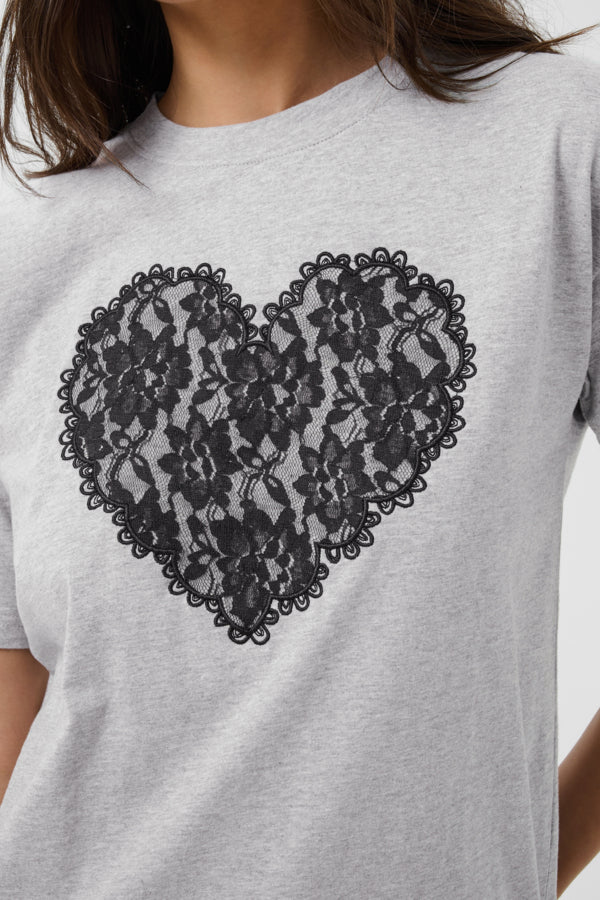 A close-up of a person wearing a light heather gray crewneck t-shirt with a large black floral lace heart graphic printed on the front.