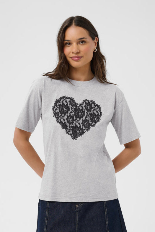 A woman modeling a light gray heather crewneck t-shirt with a black heart-shaped sequin graphic on the front, paired with a dark denim skirt, against a plain light gray background.