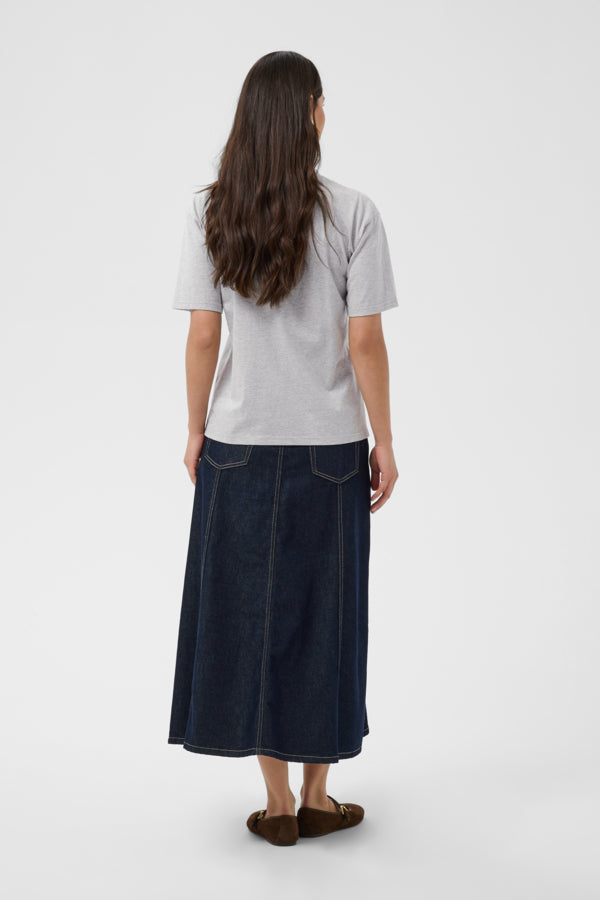 A woman shown from the back wearing a light gray t-shirt, a dark blue denim midi skirt with front patch pockets, and brown suede loafers, set against a plain light gray studio background.