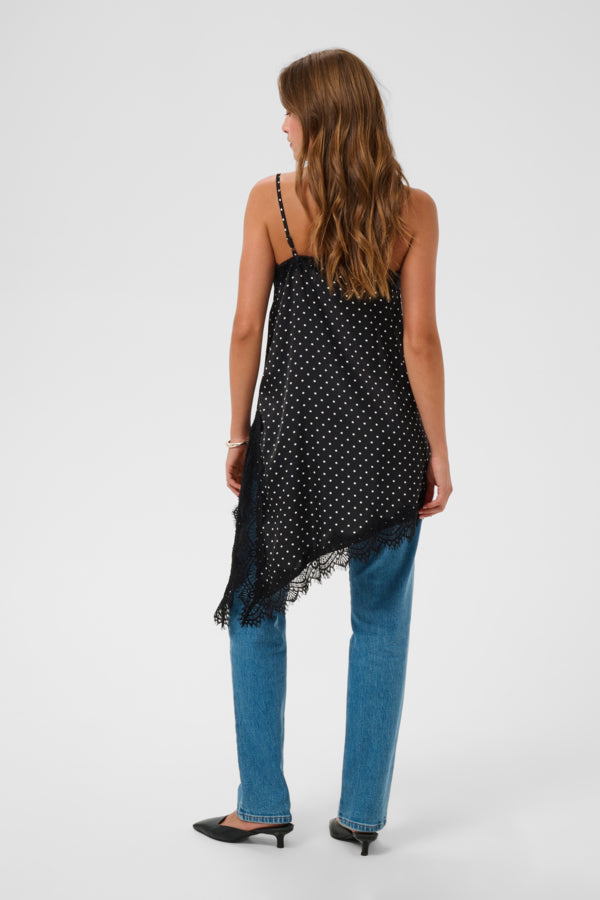 A woman shown from the back wearing a black spaghetti-strap top with white polka dots and a fringed hem, paired with blue jeans and black pointed-toe heels, against a light gray studio background.