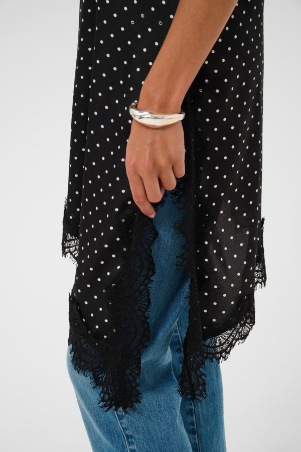 A close-up of a person wearing a black sheer long-sleeve top with white polka dots and black lace trim, paired with blue jeans and a silver cuff bracelet, against a light gray background.