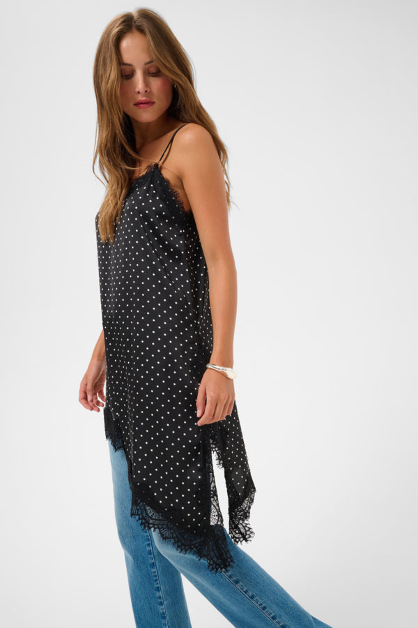 A woman modeling a black spaghetti-strap camisole top with white polka dots and a fringed hem, paired with blue jeans, against a plain light gray studio background.