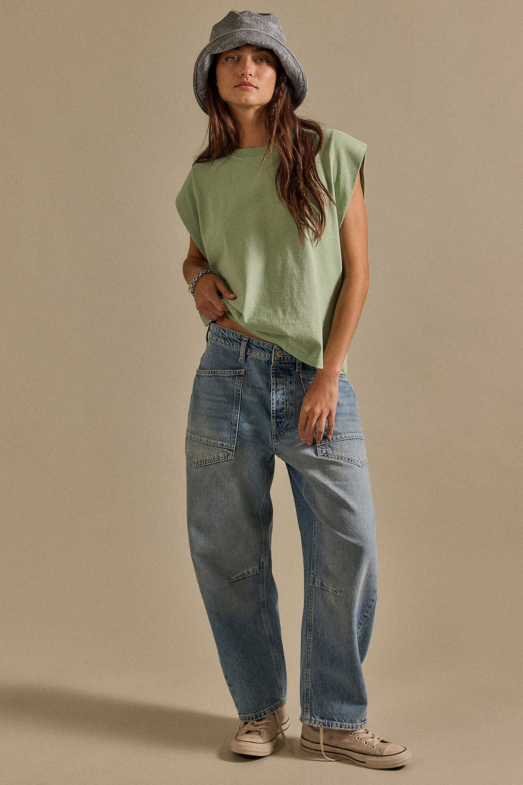 Person wearing a green sleeveless top and blue jeans with a gray bucket hat on a beige background