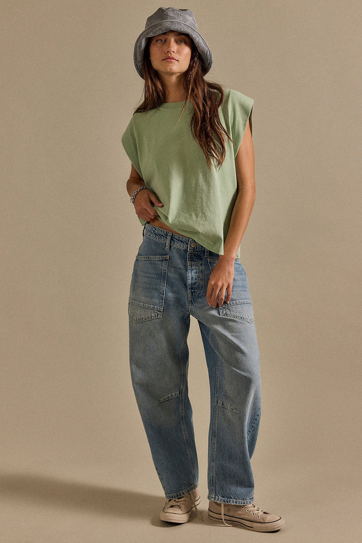 Person wearing a green sleeveless top and blue jeans with a gray bucket hat on a beige background