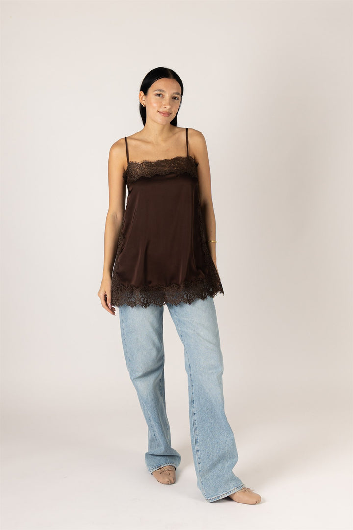 A woman modeling a dark brown sheer camisole top with lace trim, paired with light-wash wide-leg jeans, against a plain light beige studio background.